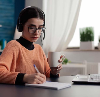 Student with headphones listening to online course and taking notes. Content creator with glasses holding cup writing on notebook. College girl with laptop studying remote at desk from home office.