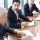 One of participants of conference consulting with older colleague while sitting by table in conference hall